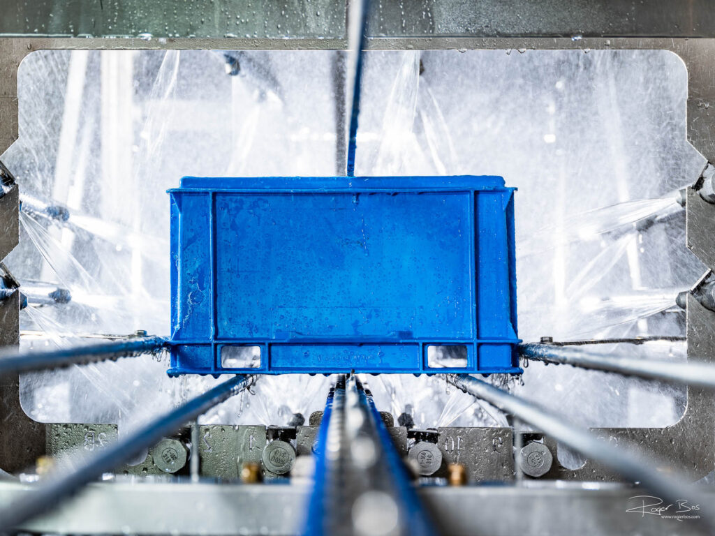 Een voedselkrat gaat door een industriële reinigings- en handlingsysteem heen. Aan alle kanten wordt er water op gespoten. Door een systeem zorgvuldig uit te lichten, ook met fotolampen in de machine, kun je de werking en techniek van de machine goed zien.