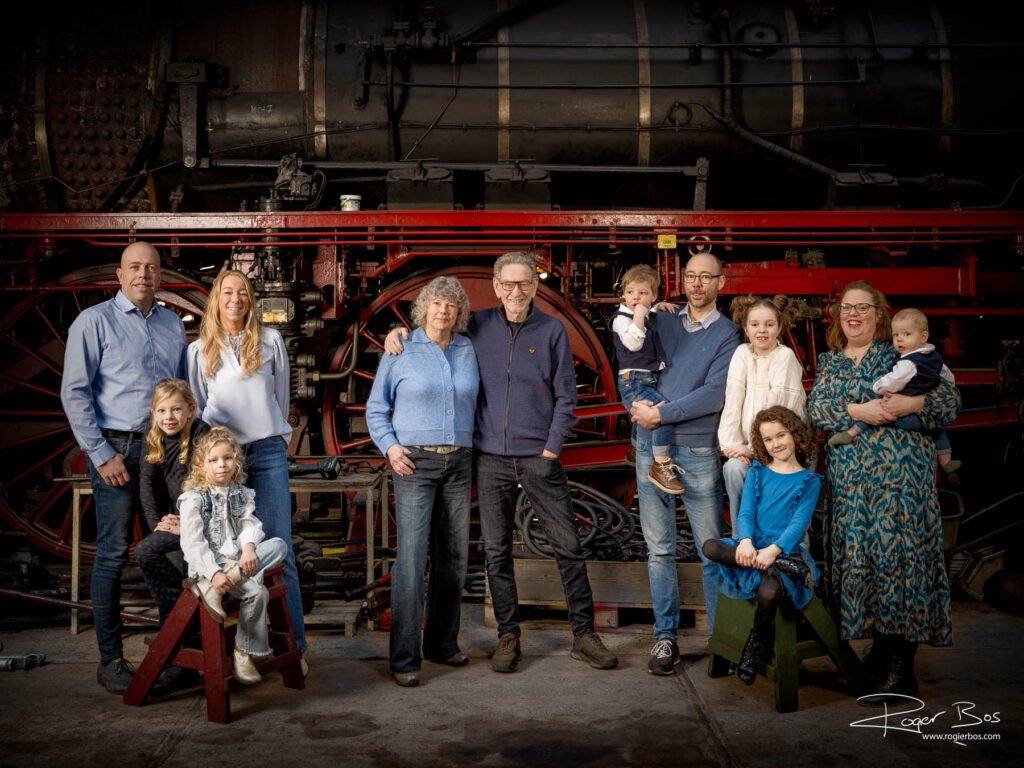 Familieportret op industriële locatie: drie generaties met stoomlocomotief op de achtergrond.