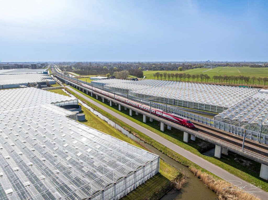 Dronefoto van Lansingerland met een hogesnelheidstrein die tussen uitgestrekte kassen rijdt