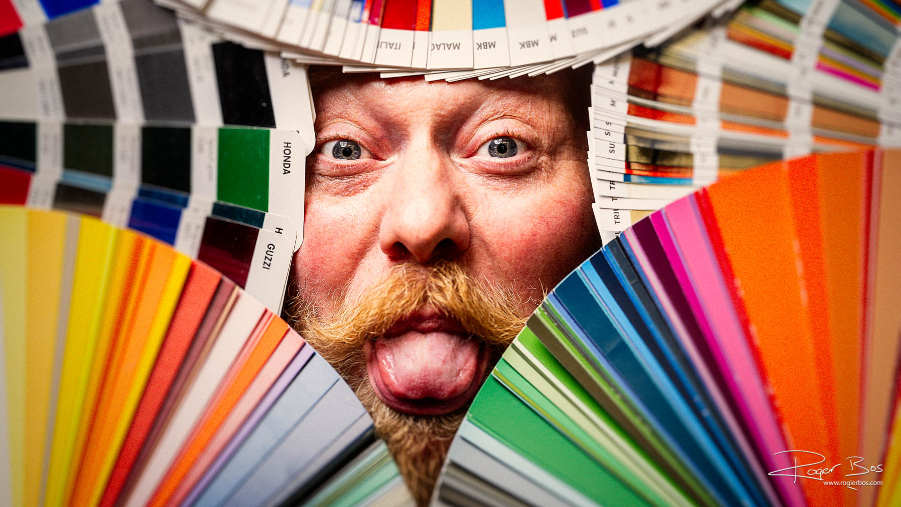 Creatieve portretfotografie met humor: kleurrijk en speels portret van een medewerker tussen verfwaaiers, gefotografeerd door portretfotograaf Rogier Bos in Nederland