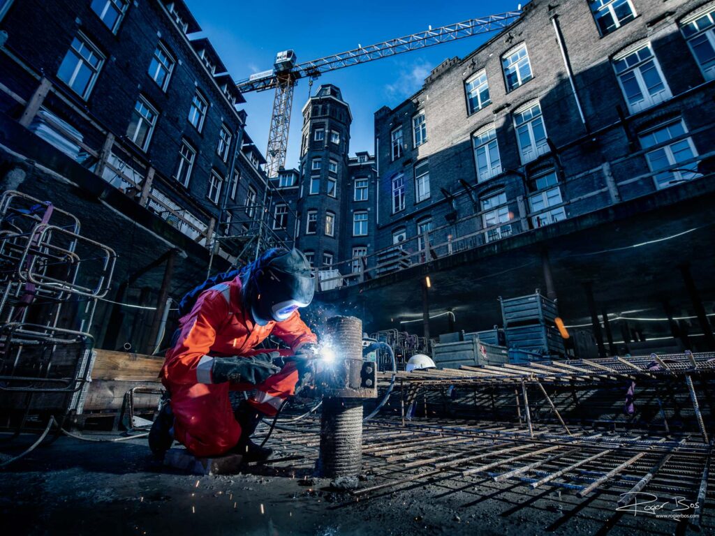 Industriële fotograaf legt lasser vast op bouwplaats in Amsterdam, werkend aan staalconstructie bij schemerlicht.