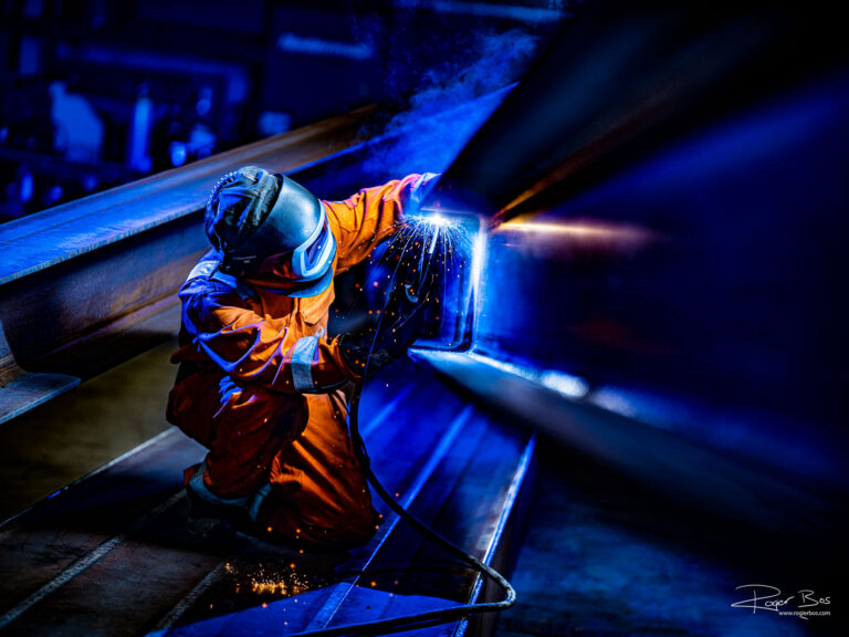 Industriële fotograaf legt MIG/MAG-lasser vast bij het lassen van zware staalconstructie in fabriekshal.