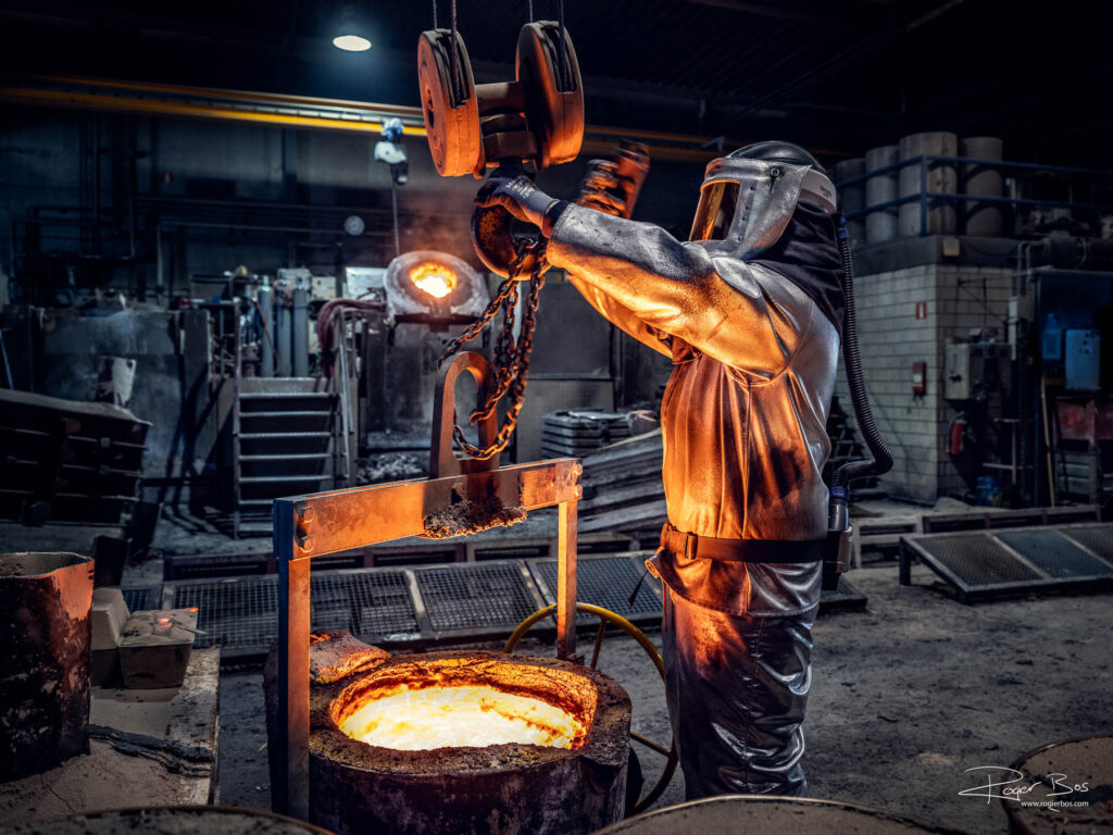 Industriële fotograaf legt smeltproces vast in metaalindustrie, met medewerker in hittebestendige kleding boven gloeiend metaal.