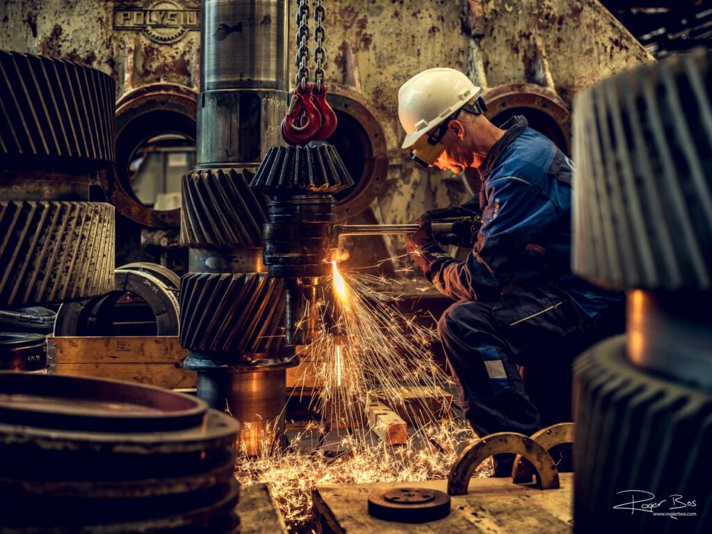 Industriële fotograaf legt lasser vast met snijbrander bij GBS International, werk aan zware tandwielen in warme lichtomstandigheden.