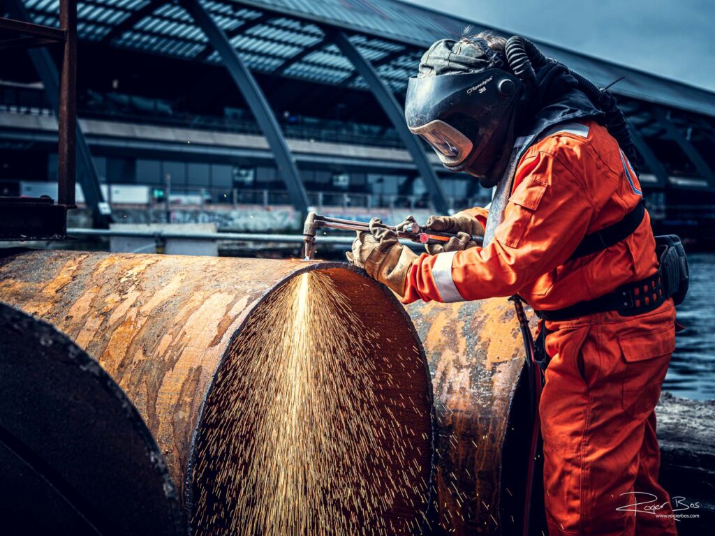 Industriële fotograaf legt branderwerker vast die stalen buis doorsnijdt met snijbrander aan het IJ bij Amsterdam Centraal.