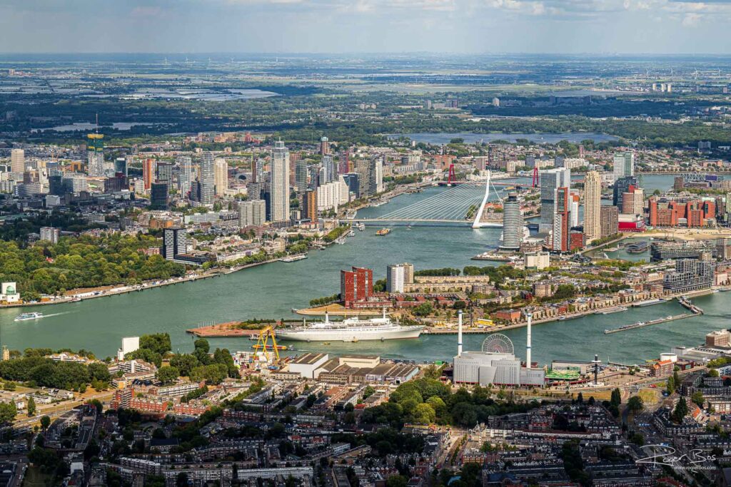 Luchtfoto van Rotterdam met de Maas, de skyline van Noord en Zuid, de Erasmusbrug en de SS Rotterdam duidelijk zichtbaar op de voorgrond.