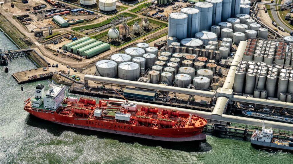 Luchtfoto van een tankterminal in Pernis met opslagtanks, laadkades en een grote rode chemicaliëntanker aan de wal.