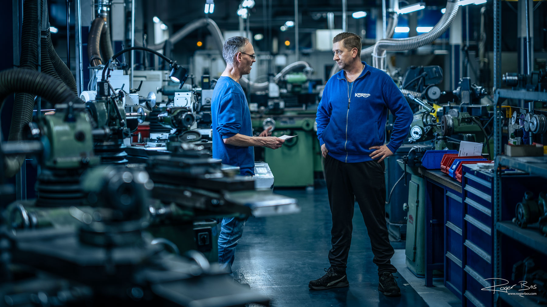 Twee technische professionals in overleg tussen machines, vastgelegd door fotograaf Rogier Bos die mens en techniek met mooie compositie, licht en nabewerking in beeld brengt.