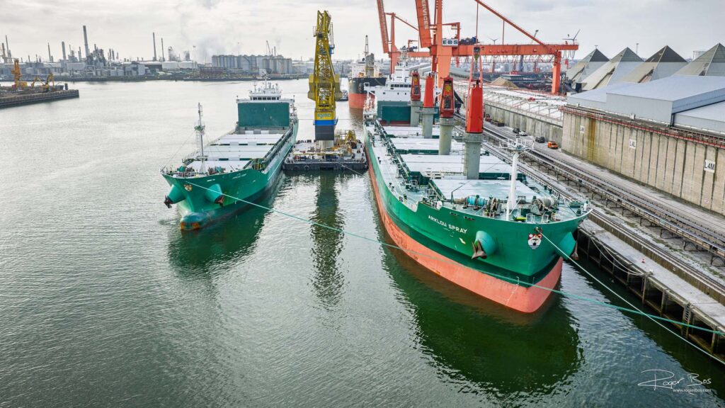 Twee groene vrachtschepen van dezelfde reder liggen naast elkaar afgemeerd in de haven van Rotterdam, met kranen en industrie op de achtergrond.