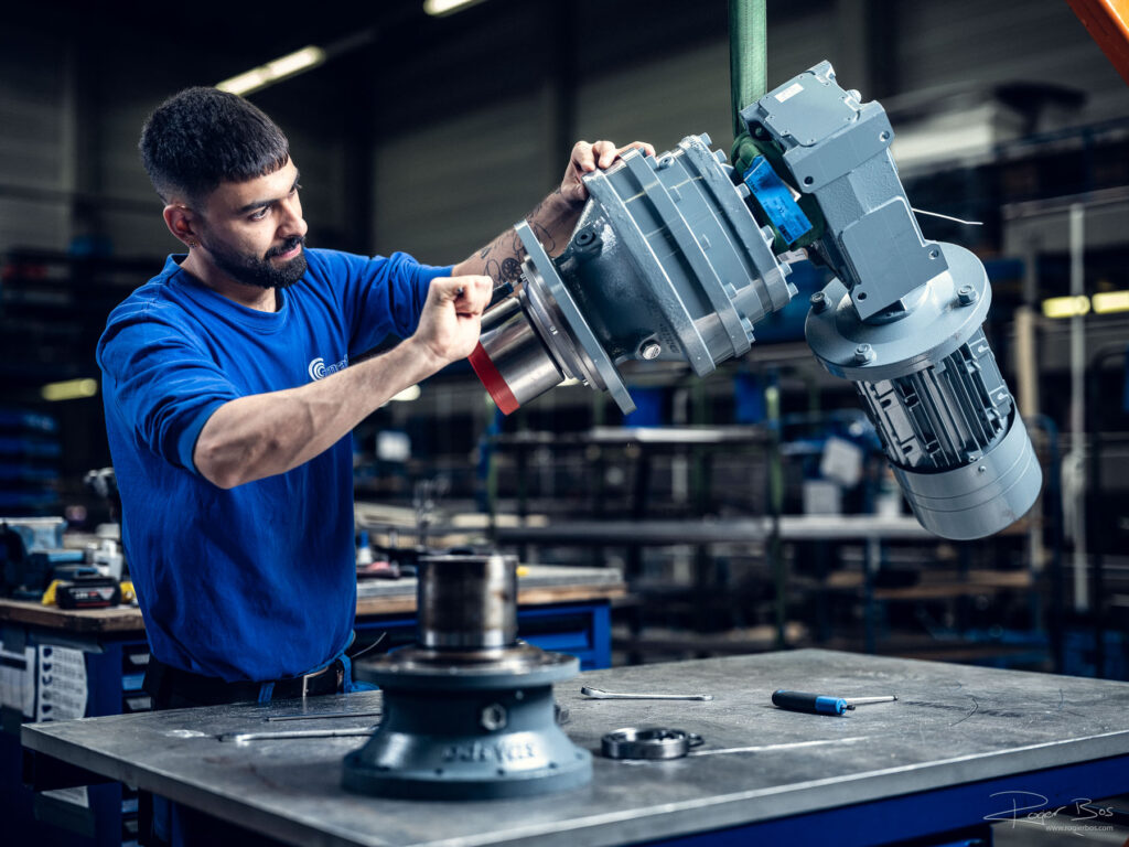 Monteur in blauwe bedrijfskleding plaatst een reductor met elektromotor op een werktafel in een industriële werkplaats