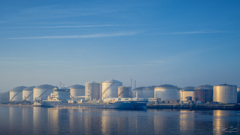 Rij grote witte opslagtanks van Vopak langs de Rotterdamse haven, met de tanker Fure Viken afgemeerd op de voorgrond in vroeg ochtendlicht.