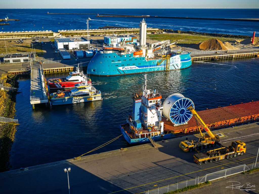 Luchtfoto van de haven van IJmuiden met een offshore windinstallatieschip, een ponton met kabeltrommel en zwaarlaadkraanwagens op de kade bij ochtendlicht