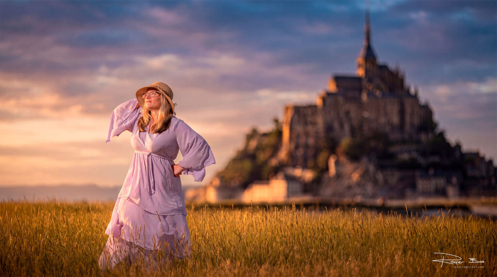 Woman in a pink dress and straw hat standing in a golden grass field at sunset with Mont Saint-Michel glowing in the background