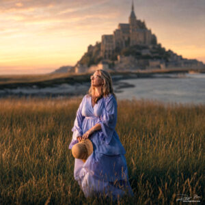 Woman in a pink dress and straw hat standing in a golden grass field at sunset with Mont Saint-Michel glowing in the background