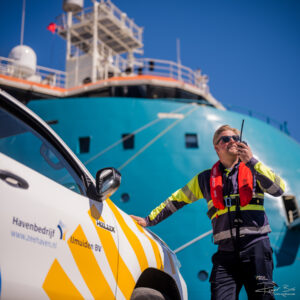 Havenmedewerker van Zeehavens IJmuiden BV met portofoon bij zijn dienstvoertuig, met de turquoise romp van de Acta Hercules op de achtergrond
